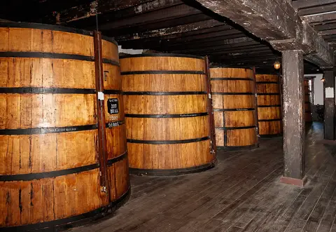 Grandes tonéis de madeira alinhados numa sala de envelhecimento de vinho Madeira, com soalho e vigas antigas