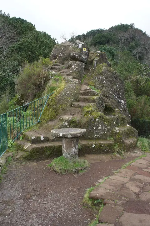 Rocha com degraus talhados e mesa de pedra junto a guarda metálica, em trilho de miradouro