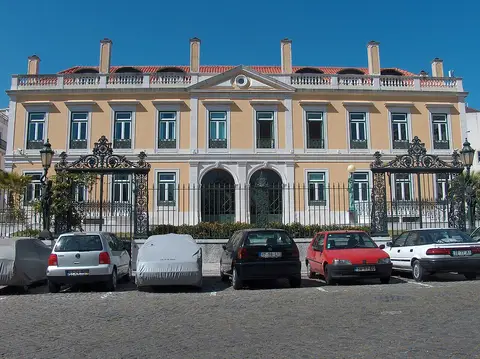 Fachada de palacete amarelo com portões de ferro forjado e carros estacionados em frente, em Lisboa