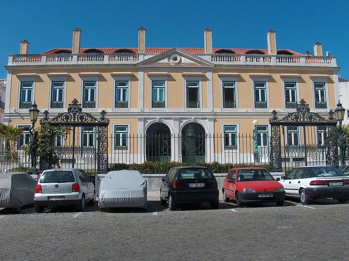 Fachada de palacete amarelo com portões de ferro forjado e carros estacionados em frente, em Lisboa