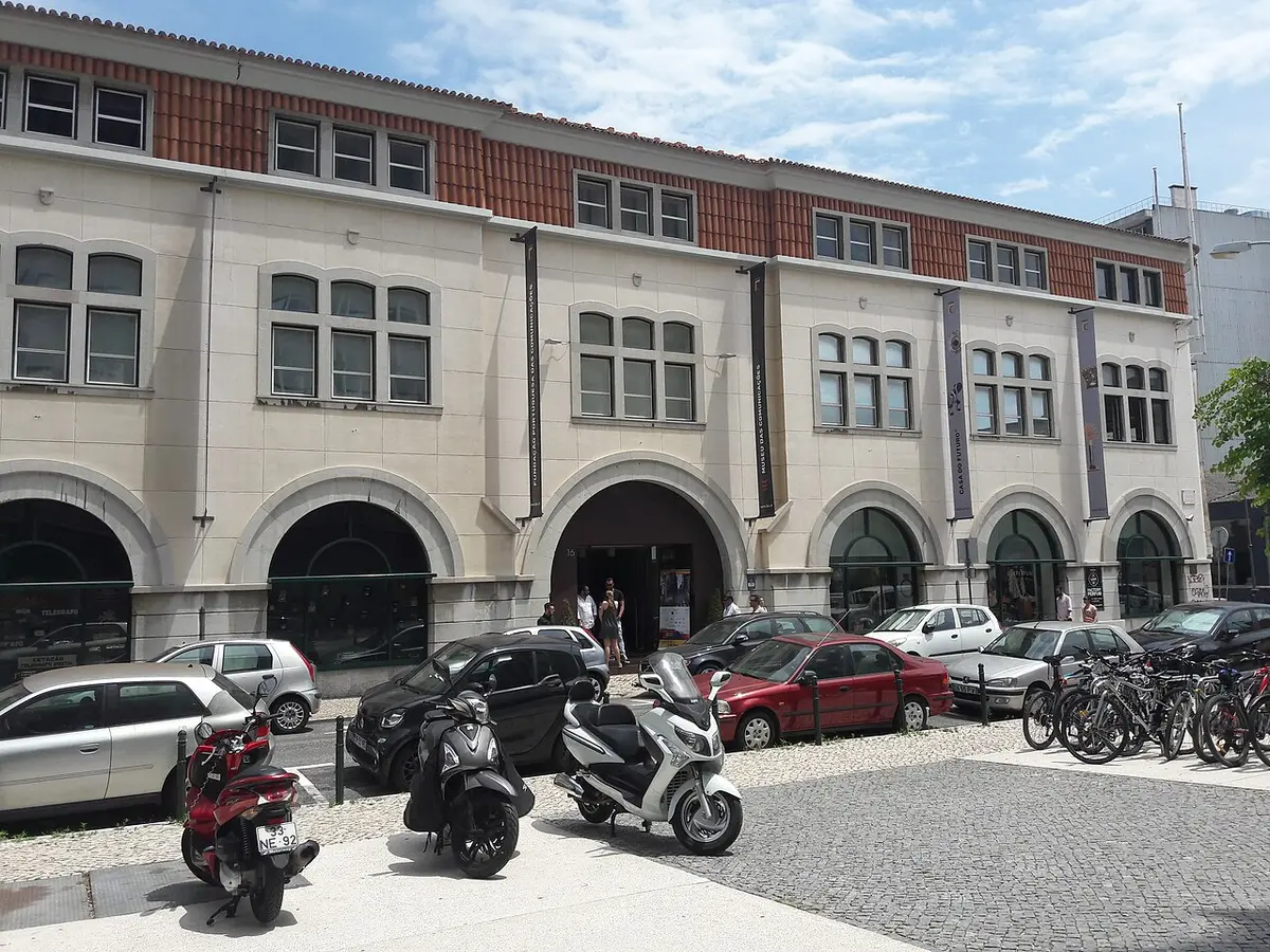 Fachada do Museu das Comunicações em Lisboa, com arcadas e veículos estacionados em frente.