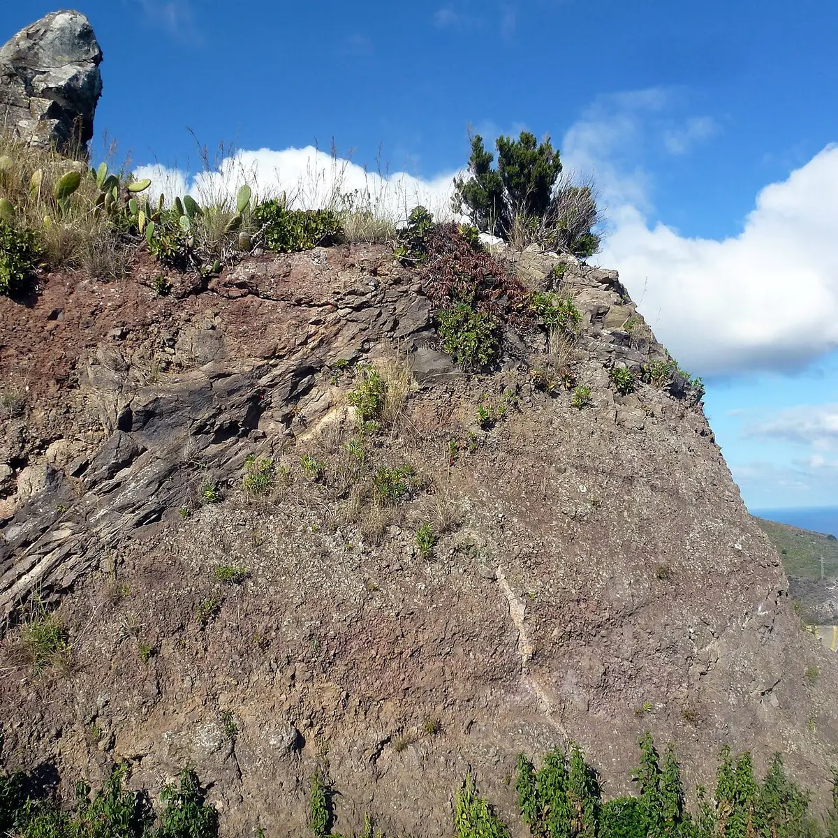 Encosta rochosa com vegetação no Pico do Facho, Machico, sob céu azul com nuvens