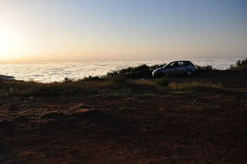 Carro estacionado em terreno de terra vermelha acima de um mar de nuvens ao pôr do sol, na Madeira