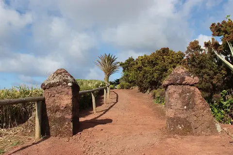 Caminho de terra vermelha entre pilares de pedra vulcânica a caminho do miradouro, com vegetação e céu nublado
