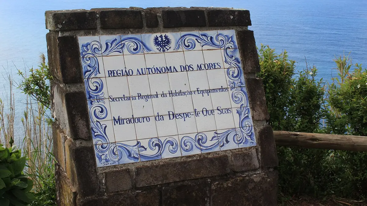 Painel de azulejos em pedra com o nome do Miradouro da Despe-te que Suas, com o mar ao fundo