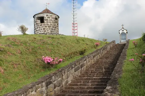 Escadaria de pedra a subir para torre de vigia das baleias em pedra, com nicho religioso e flores.