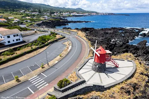Moinho de vento vermelho junto à costa de lava e à estrada marginal, com vista para o mar
