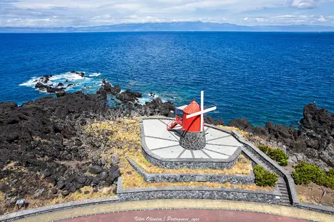 Moinho de vento vermelho sobre plataforma de pedra junto à costa rochosa, com o mar azul em fundo