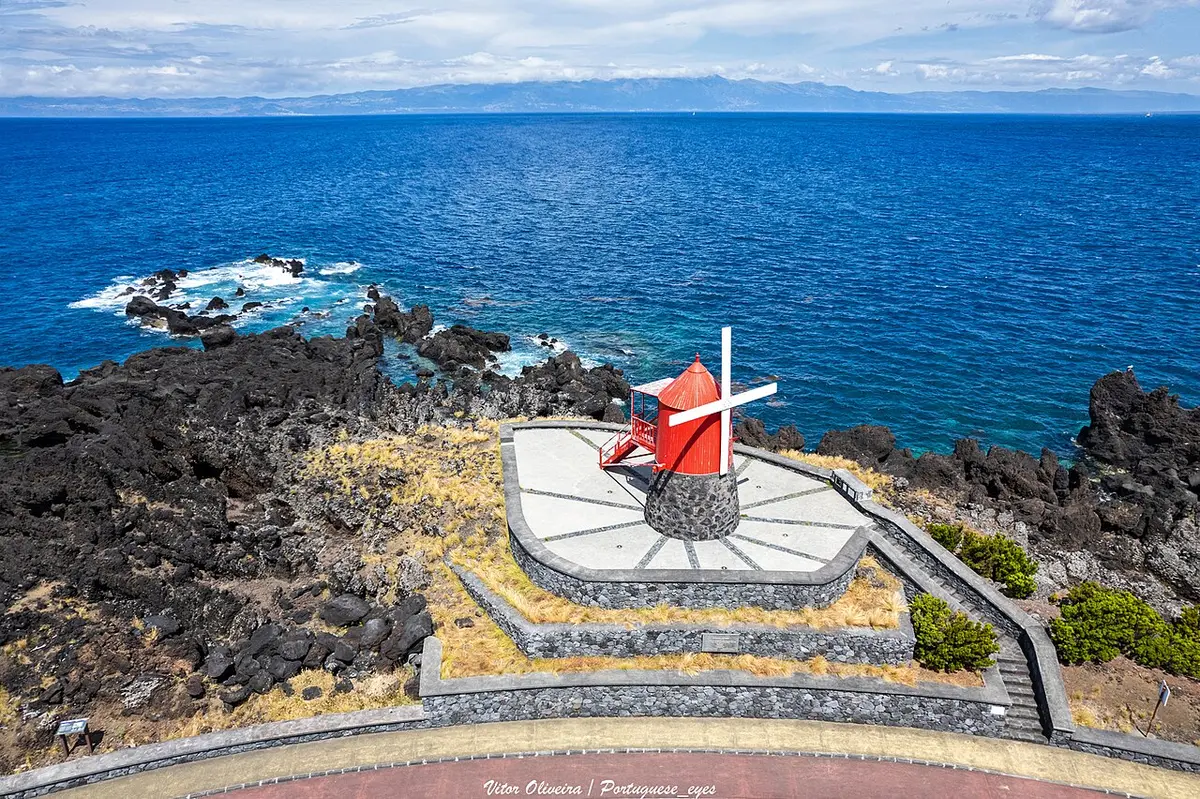 Moinho de vento vermelho sobre plataforma de pedra junto à costa rochosa, com o mar azul em fundo