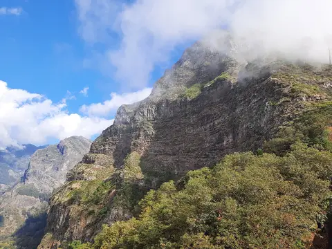 Encostas rochosas e escarpadas do Curral das Freiras envoltas em nuvens, com vegetação em primeiro plano.
