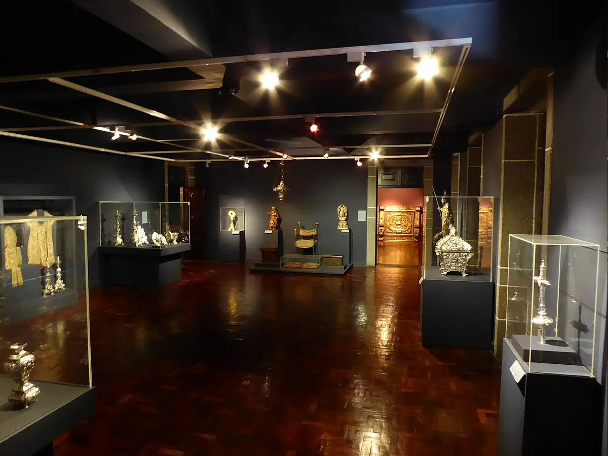 Sala de museu de arte sacra com vitrinas de peças de ourivesaria e imagens religiosas douradas