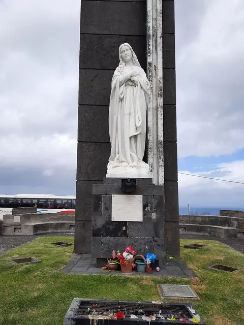 Monumento com estátua de Nossa Senhora da Conceição em pedra escura, com flores na base, no miradouro