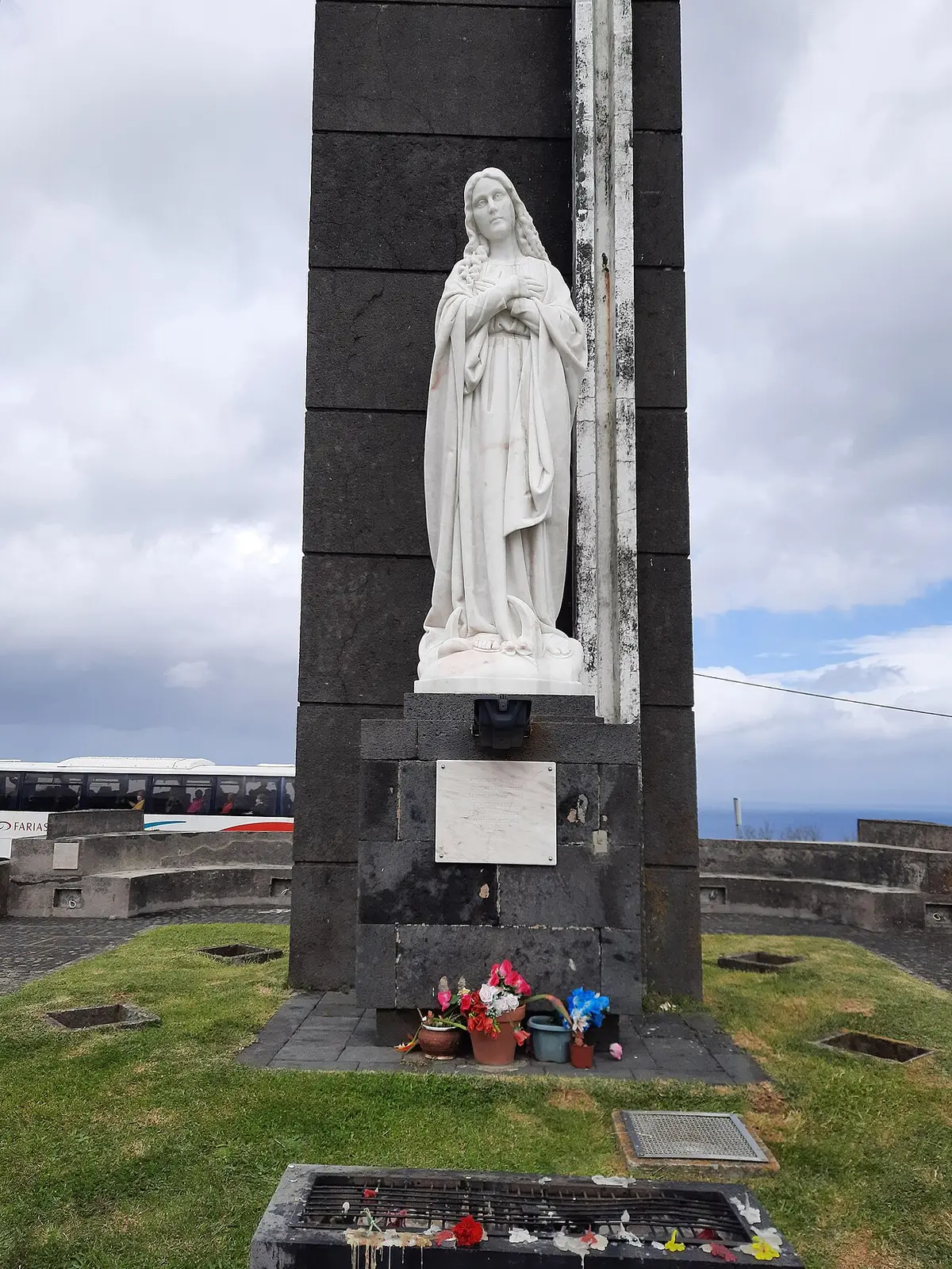 Monumento com estátua de Nossa Senhora da Conceição em pedra escura, com flores na base, no miradouro