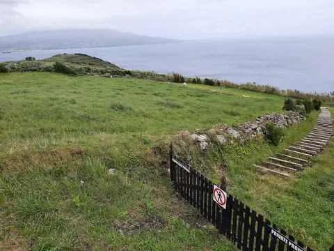 Vista sobre o mar e a costa a partir do miradouro, com prado verde, escadas e vedação com sinal de propriedade privada