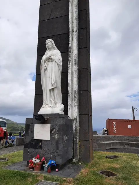 Estátua branca de Nossa Senhora junto a monumento de pedra escura no miradouro, com flores na base.