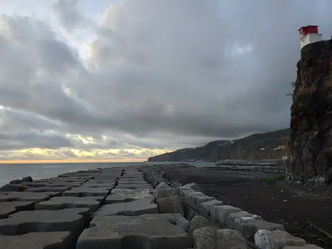 Farol no topo de uma rocha junto ao mar, com quebra-mar de blocos de betão e céu nublado.