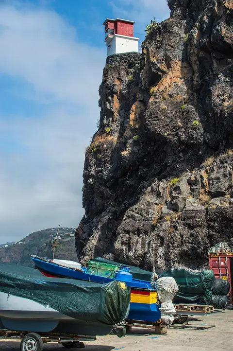 Farol branco e vermelho no topo de uma falésia, com barcos de pesca cobertos em terra