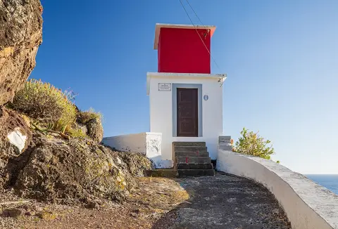 Pequeno farol branco com topo vermelho junto à falésia, com o mar ao fundo, na Ribeira Brava