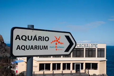 Placa indicativa do Aquário da Madeira em Porto Moniz, com edifício e mar ao fundo