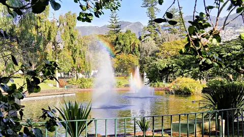 Lago com repuxos e arco-íris no Parque de Santa Catarina, Funchal, rodeado de árvores e montanhas ao fundo