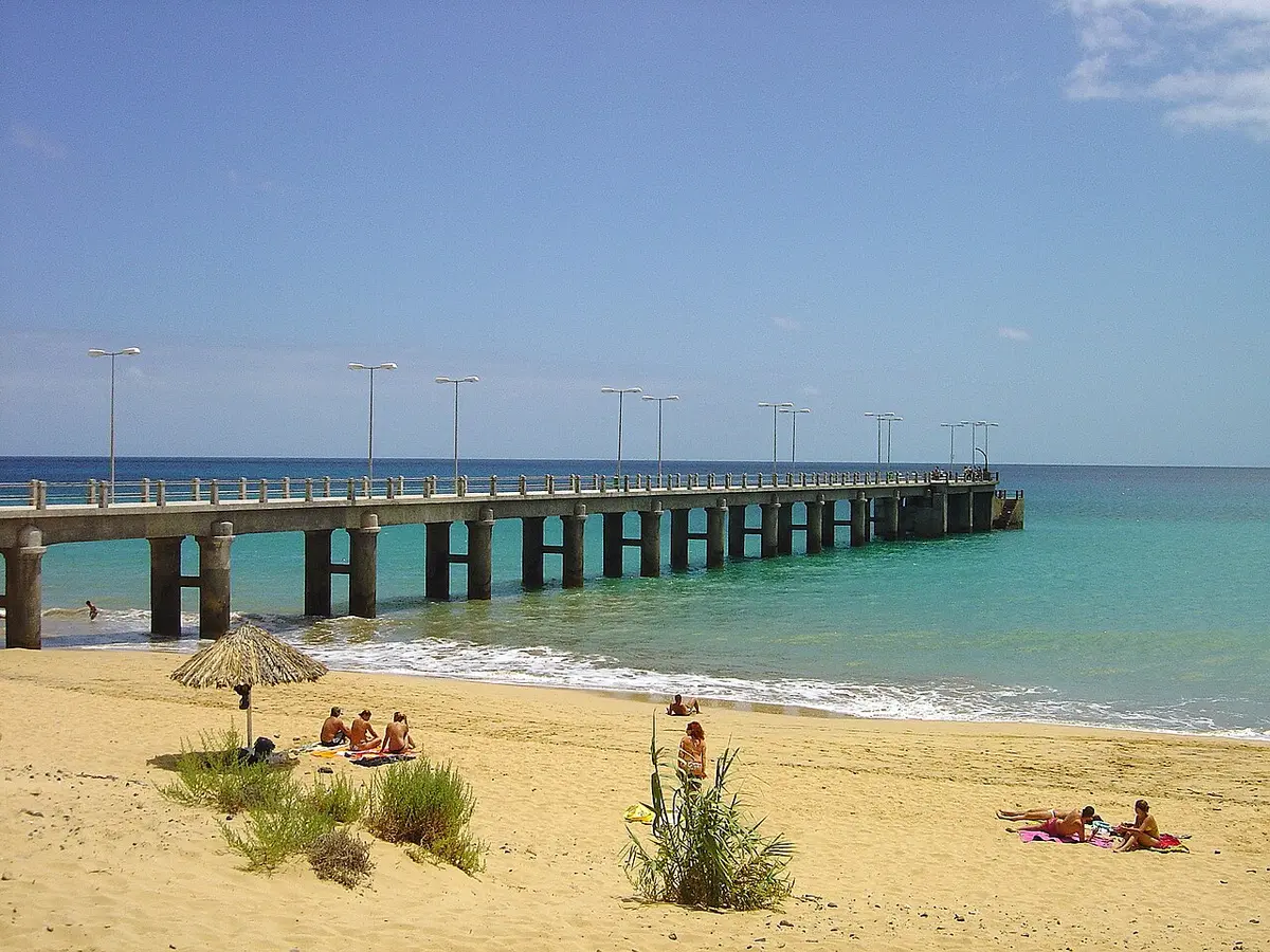 Pontão de betão sobre mar azul-turquesa, visto da praia de areia dourada do Porto Santo