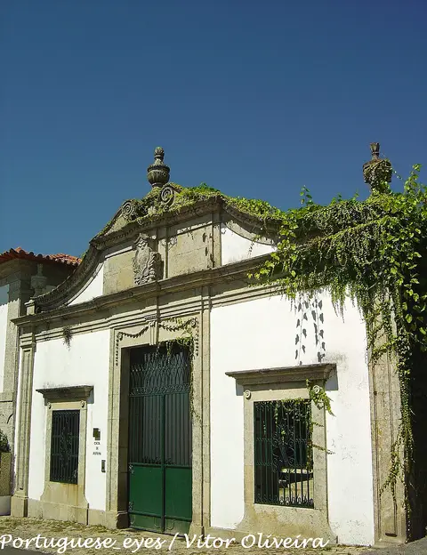 Fachada branca da Casa de Alfena em Travassos, com portão de ferro verde, brasão e vegetação sobre o muro.