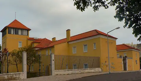Edifício amarelo com telhado de telha e torre envidraçada, rodeado por gradeamento, vista da rua