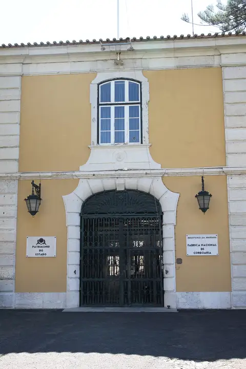 Entrada principal da Fábrica Nacional da Cordoaria, com portão de ferro e placas identificativas.