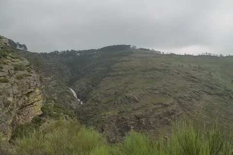 Vista do vale com a queda de água das Fisgas de Ermelo entre encostas rochosas e céu nublado