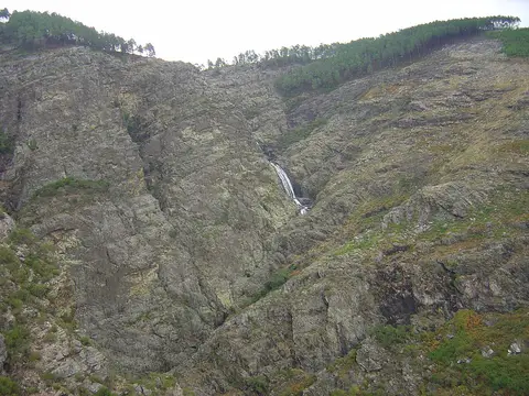 Queda de água das Fisgas de Ermelo a descer uma encosta rochosa com vegetação e pinhal no topo