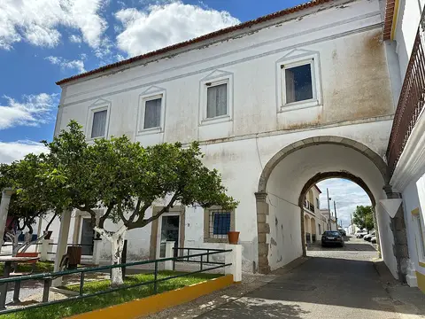Edifício branco da Casa do Arco, em Vila de Frades, com arco sobre a rua e árvores em frente.