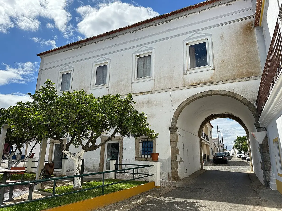 Edifício branco da Casa do Arco, em Vila de Frades, com arco sobre a rua e árvores em frente.