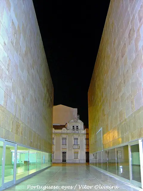 Passagem entre os volumes de pedra do Centro de Artes de Sines à noite, com edifício antigo ao fundo