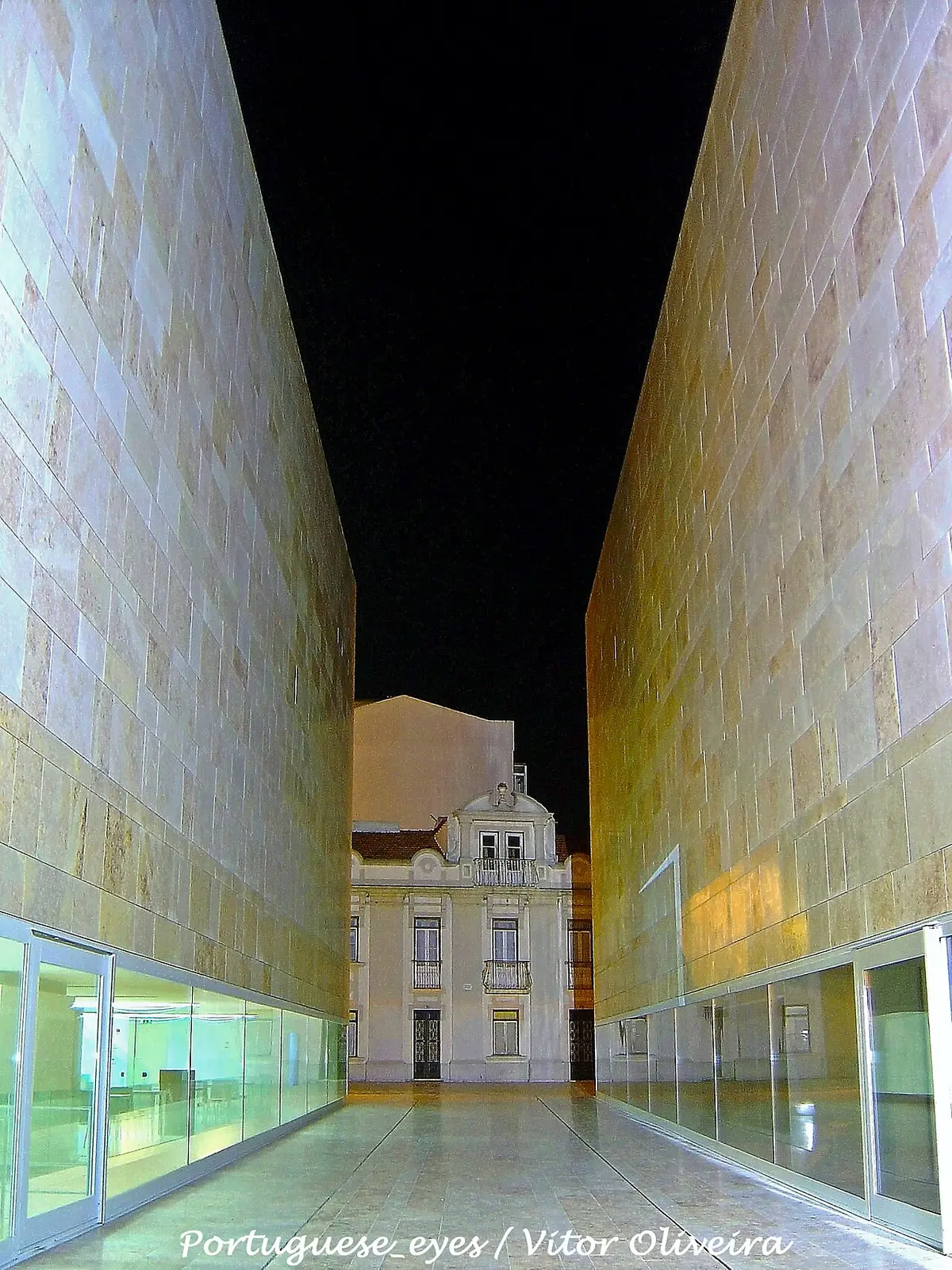 Passagem entre os volumes de pedra do Centro de Artes de Sines à noite, com edifício antigo ao fundo