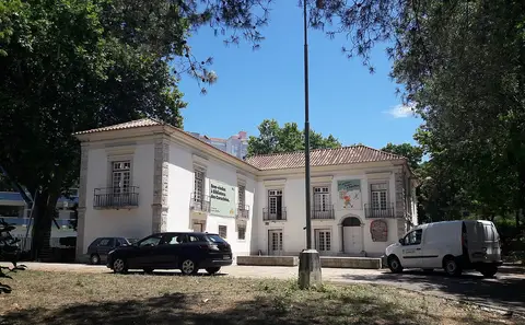 Edifício branco da Biblioteca dos Coruchéus, em Lisboa, rodeado de árvores e com carros estacionados