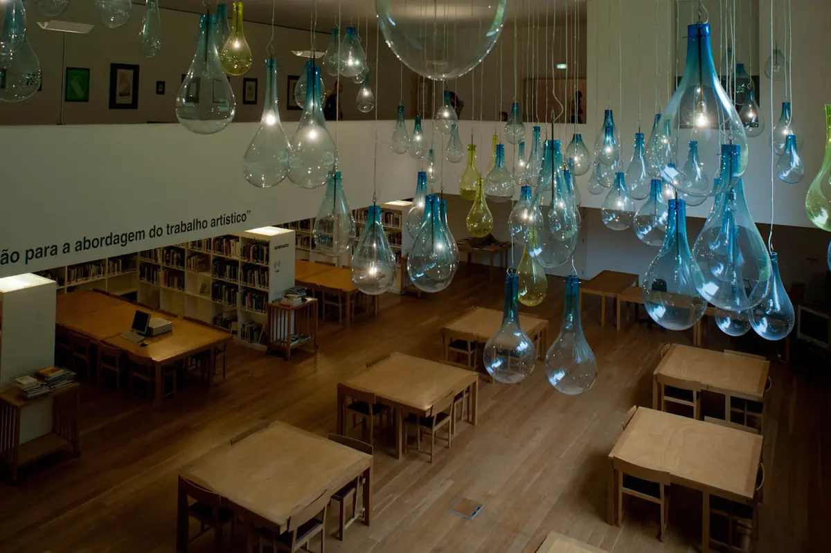 Interior da Biblioteca de Serralves com candeeiros de vidro suspensos, mesas de madeira e estantes de livros.