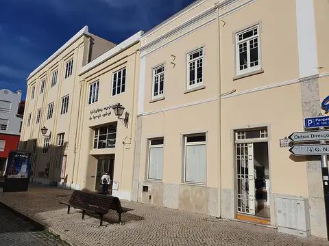 Fachada da Biblioteca Municipal de Torres Vedras, edifício claro com banco de madeira na calçada.