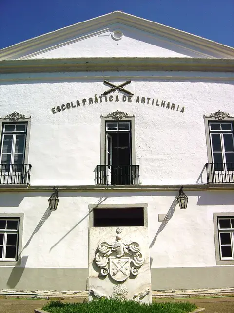 Fachada branca da Escola Prática de Artilharia, com letreiro, brasão em pedra e varandas de ferro.