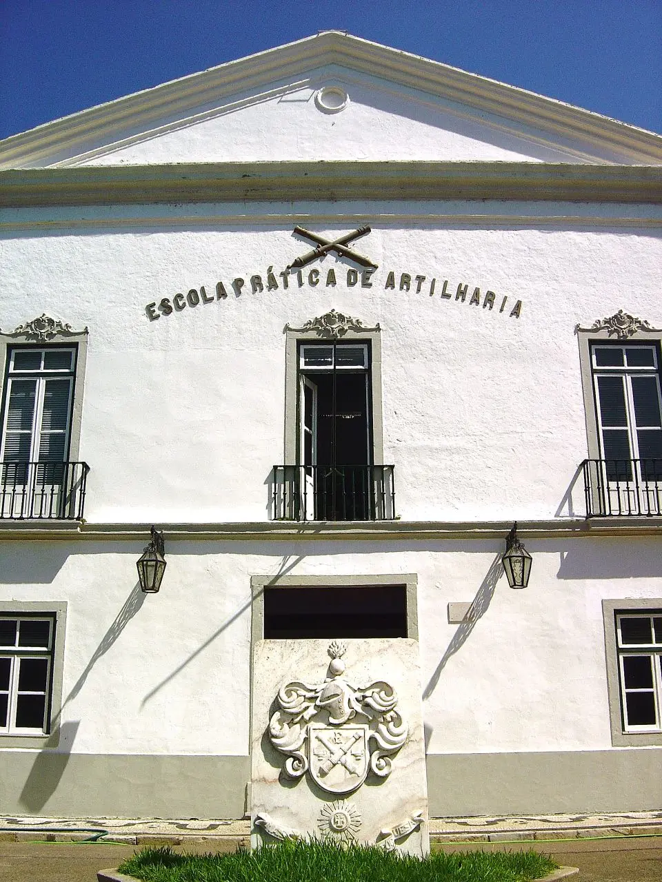 Fachada branca da Escola Prática de Artilharia, com letreiro, brasão em pedra e varandas de ferro.