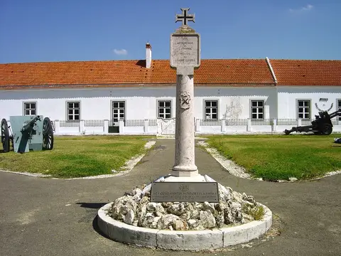 Monumento com cruz em frente ao edifício branco do Museu da Escola Prática de Artilharia, com canhões antigos.