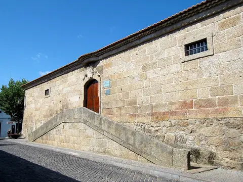 Edifício histórico em cantaria de granito com portada em arco e escadaria exterior, sob céu azul.