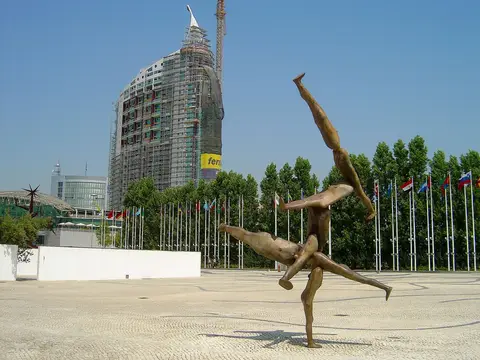 Escultura de bronze numa praça do Parque das Nações, com bandeiras e torre em construção ao fundo