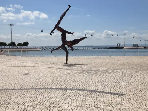 Escultura de figuras humanas em bronze no passeio calcetado do Parque das Nações, com teleférico e rio ao fundo