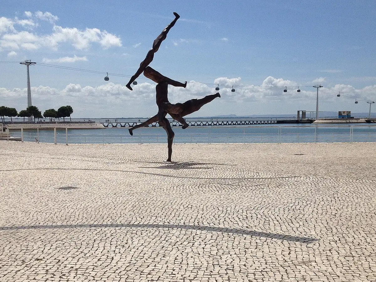 Escultura de figuras humanas em bronze no passeio calcetado do Parque das Nações, com teleférico e rio ao fundo