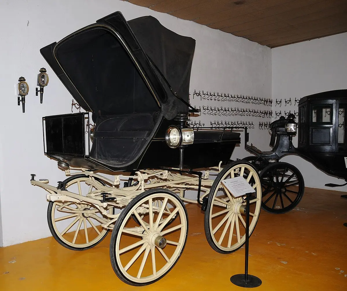 Carruagem antiga tipo faetonte com rodas claras exposta numa sala de museu de carros de cavalos