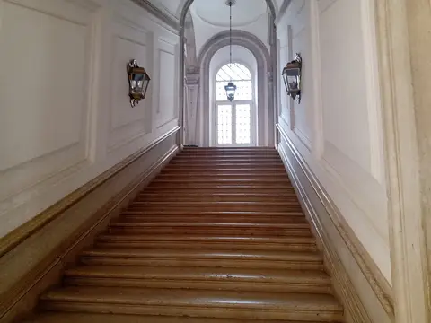 Escadaria monumental em mármore num palácio, com lanternas nas paredes e janela em arco ao fundo