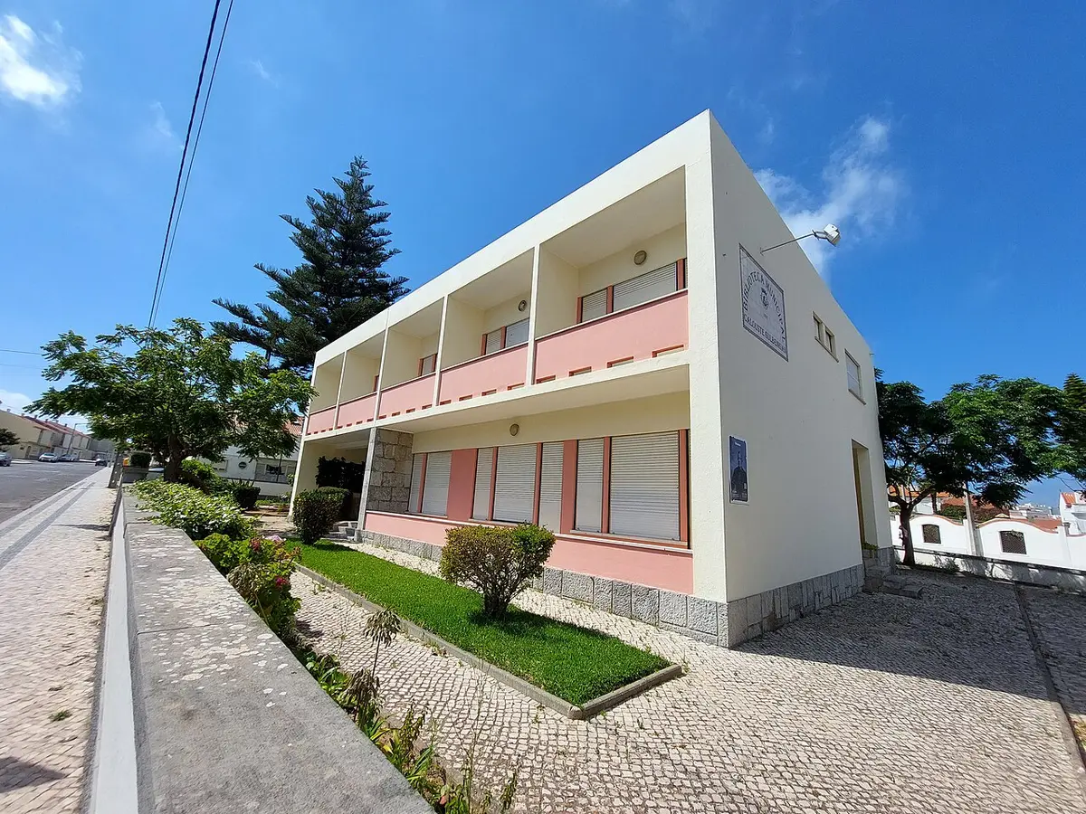 Edifício branco e rosa da Biblioteca Municipal de Peniche, com jardim e calçada em frente.