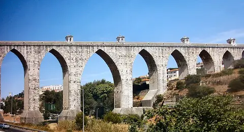 Arcos de pedra do Aqueduto das Águas Livres sobre o vale, em Lisboa, com céu azul.