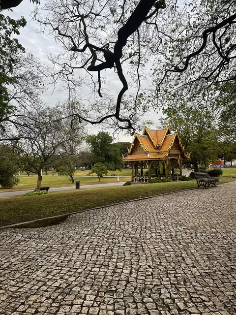 Pavilhão tailandês dourado num jardim relvado, com caminho em calçada portuguesa e árvores em redor.