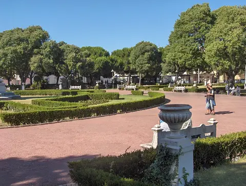 Jardim com sebes recortadas, árvores frondosas e caminho de saibro avermelhado junto a vaso decorativo em pedra.